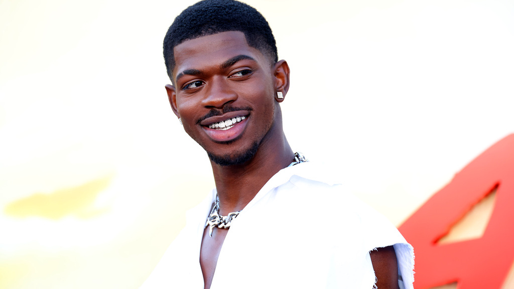 Lil Nas X sonriendo, con dientes con brillantes, camisa blanca sin mangas y cadena plateada, en una alfombra roja con fondo amarillo y rojo
