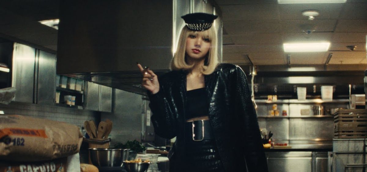 Fotograma del video oficial de «Tarantino»: LISA, con peluca rubia carré, gorra negra de visera y saco de cuero negro, parada en la cocina industrial de un hotel