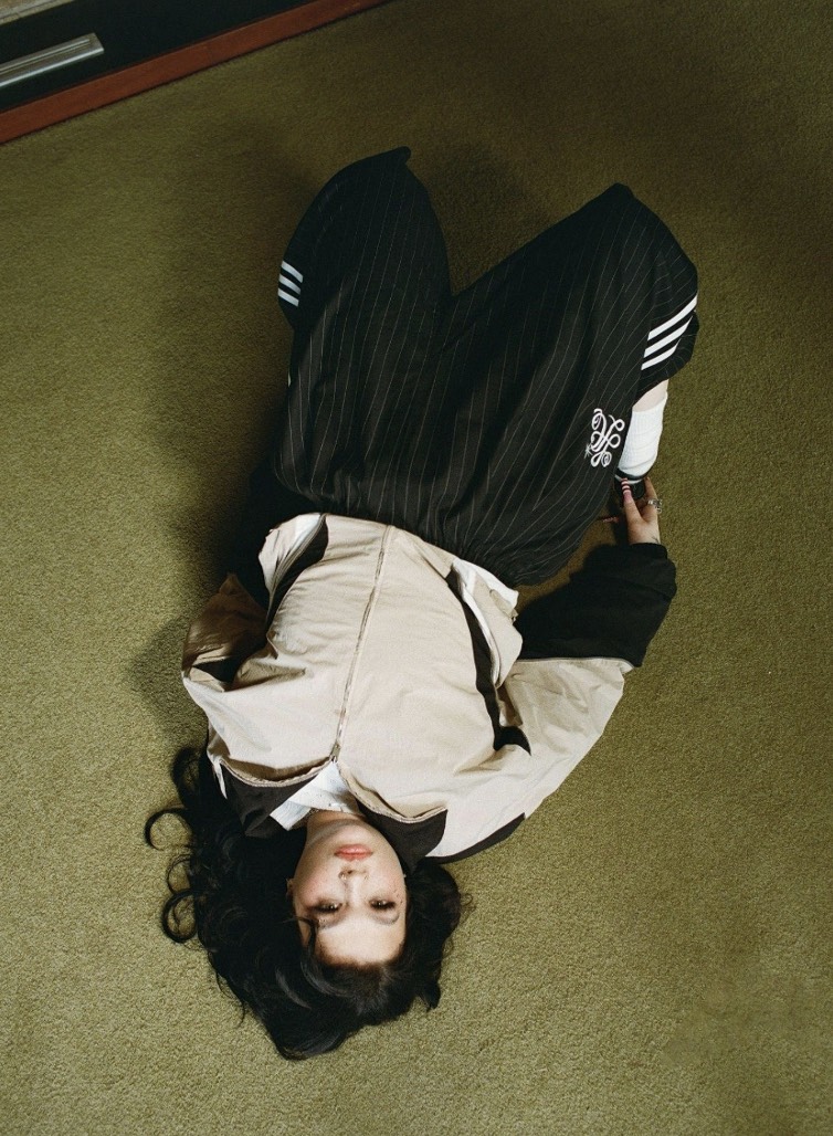 Foto de prensa de Lola Young acostada boca arriba sobre una alfombra verde, vista desde arriba, con campera beige y negra y pantalón negro a rayas finas