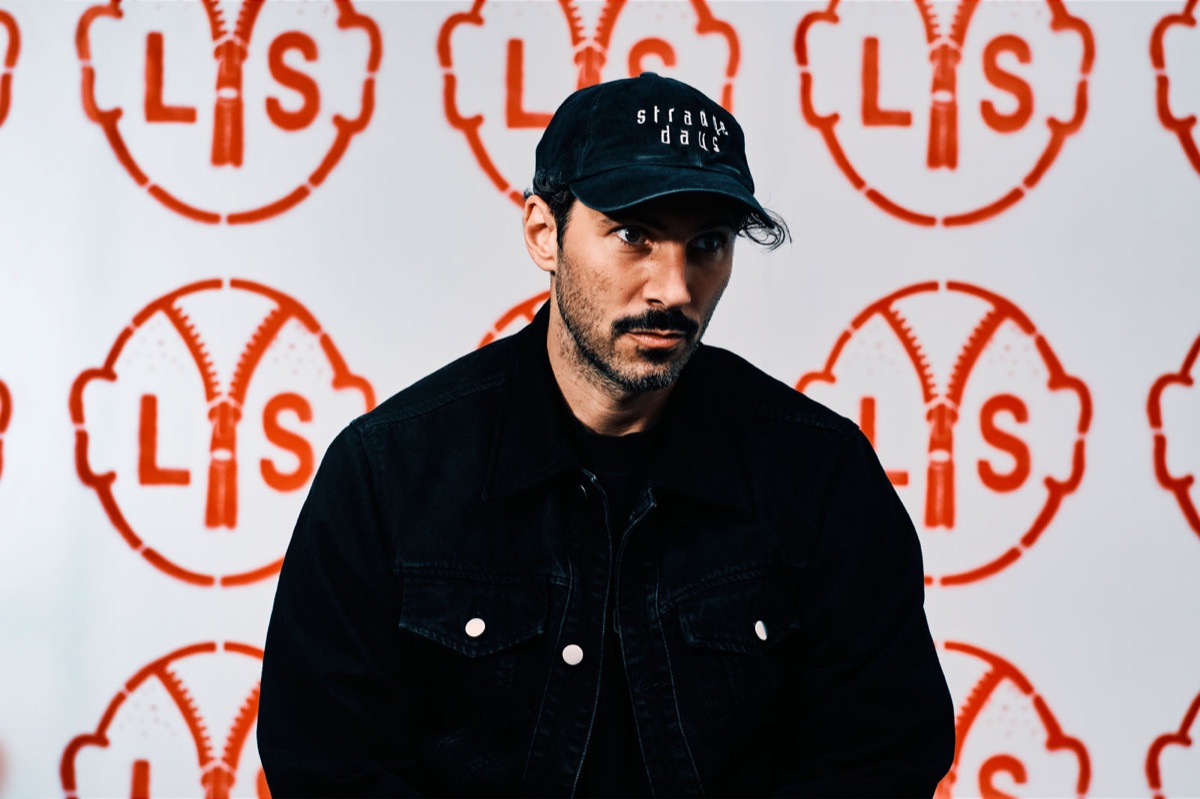 Foto de prensa oficial de Lorenzo Senni para "All Ages", por Jim C. Nedd: el productor, con gorra y campera de jean negras, sentado frente a una pared llena del logo rojo del disco