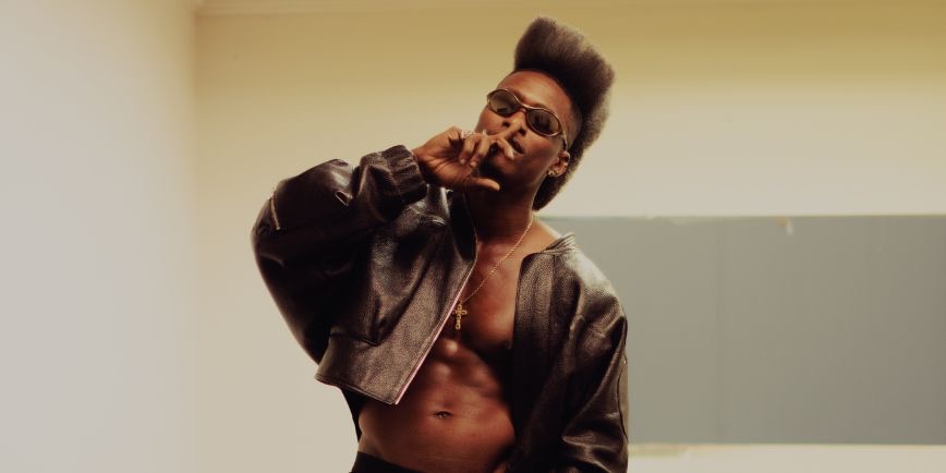 Foto de prensa de Lucky Daye con campera de cuero abierta, anteojos y peinado alto, frente a una pared clara