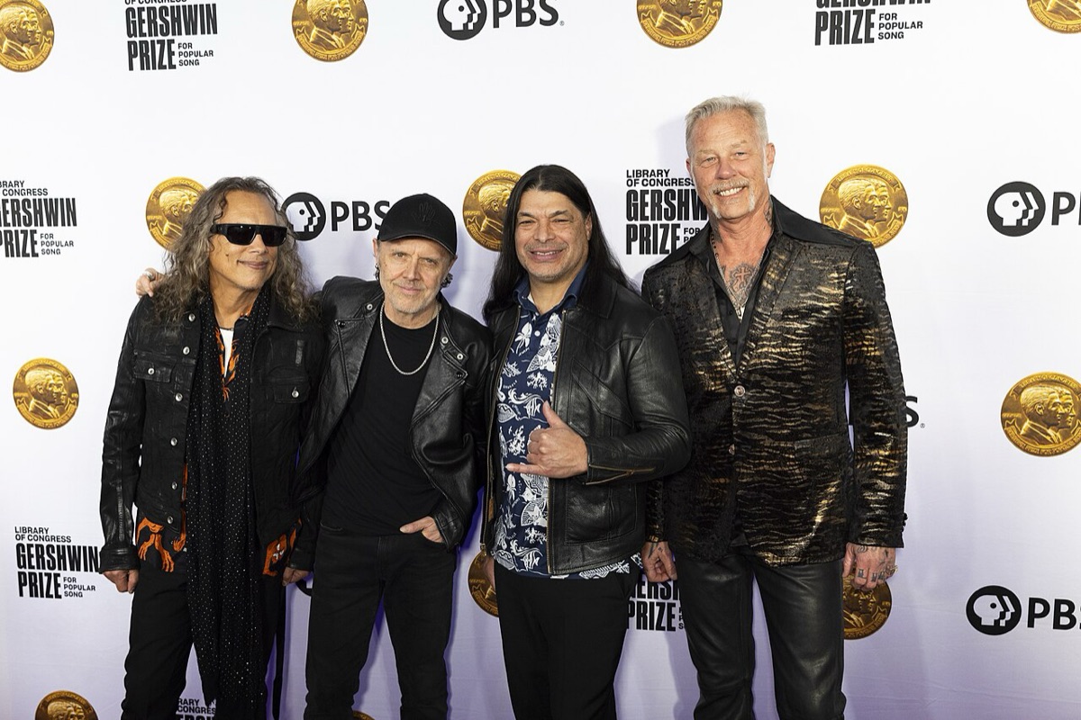 Foto de archivo de Metallica: Kirk Hammett con anteojos negros, Lars Ulrich con gorra, Robert Trujillo con camisa estampada y James Hetfield con saco dorado y negro, posando abrazados frente a un panel blanco del Premio Gershwin en 2024