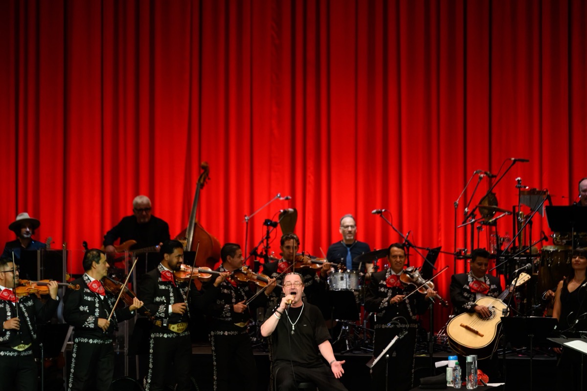 Mike Patton canta sentado al frente del escenario del Palacio de Bellas Artes, rodeado por los músicos del Mariachi Gama 1000 con trajes de charro negros y moños rojos, frente a un telón rojo