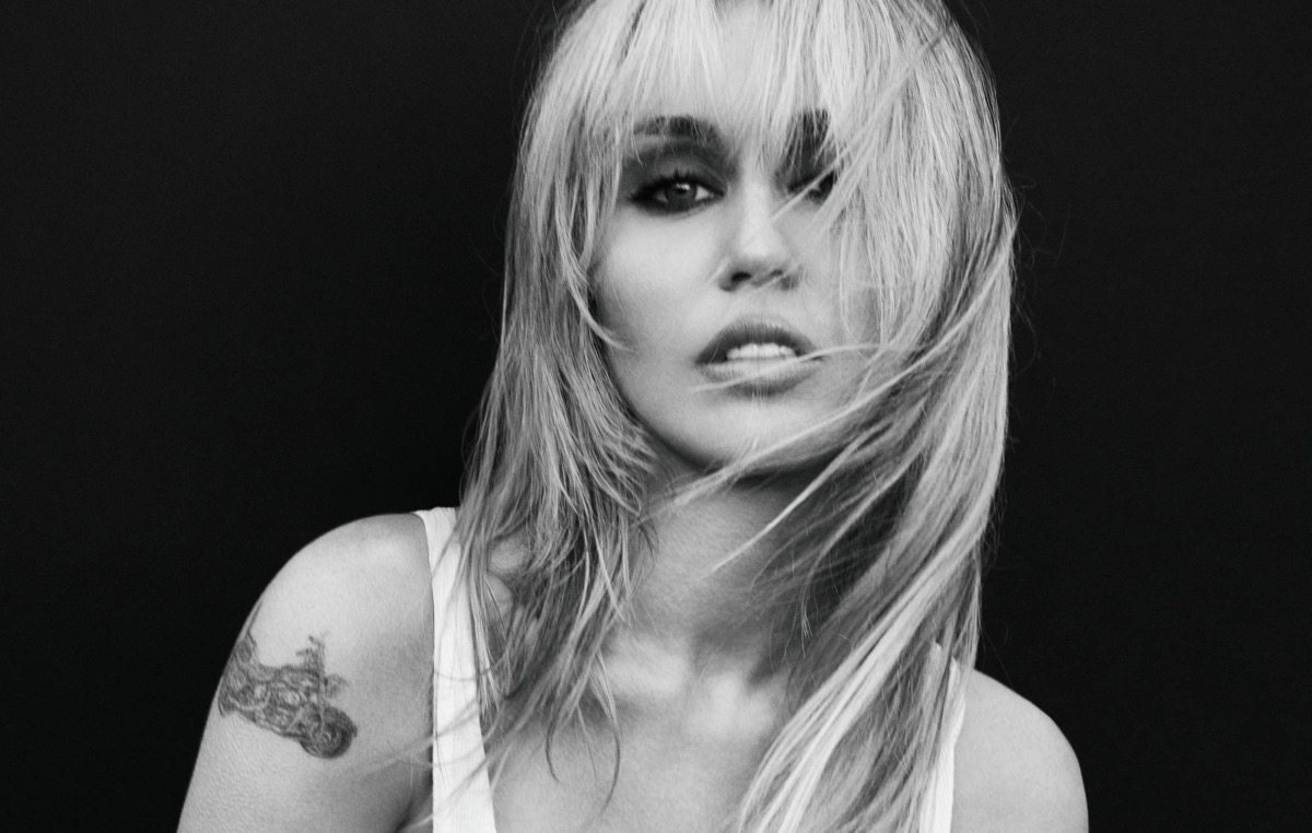 Foto de prensa oficial de Miley Cyrus para la etapa de "Bass Persuades", en blanco y negro: con flequillo y el pelo rubio largo cruzándole la cara, musculosa blanca y un tatuaje en el brazo derecho, mira a cámara sobre un fondo negro. Foto: Mert Alas