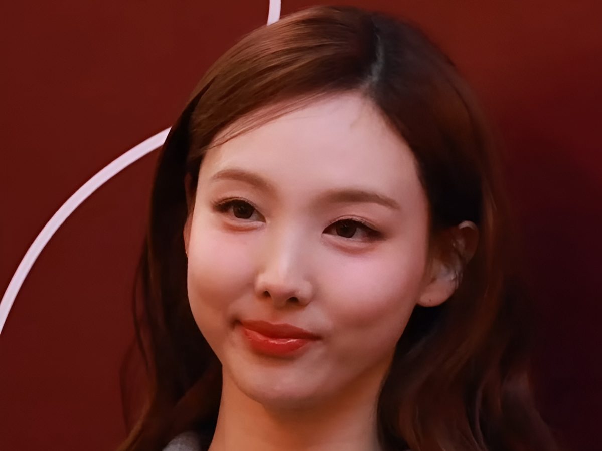 Nayeon de TWICE sonriendo en un evento de Maison Kitsuné en octubre de 2024, con pelo castaño suelto y campera gris con cierre, frente a un fondo rojo oscuro (foto de archivo). Foto: TV10, CC BY 3.0, vía Wikimedia Commons