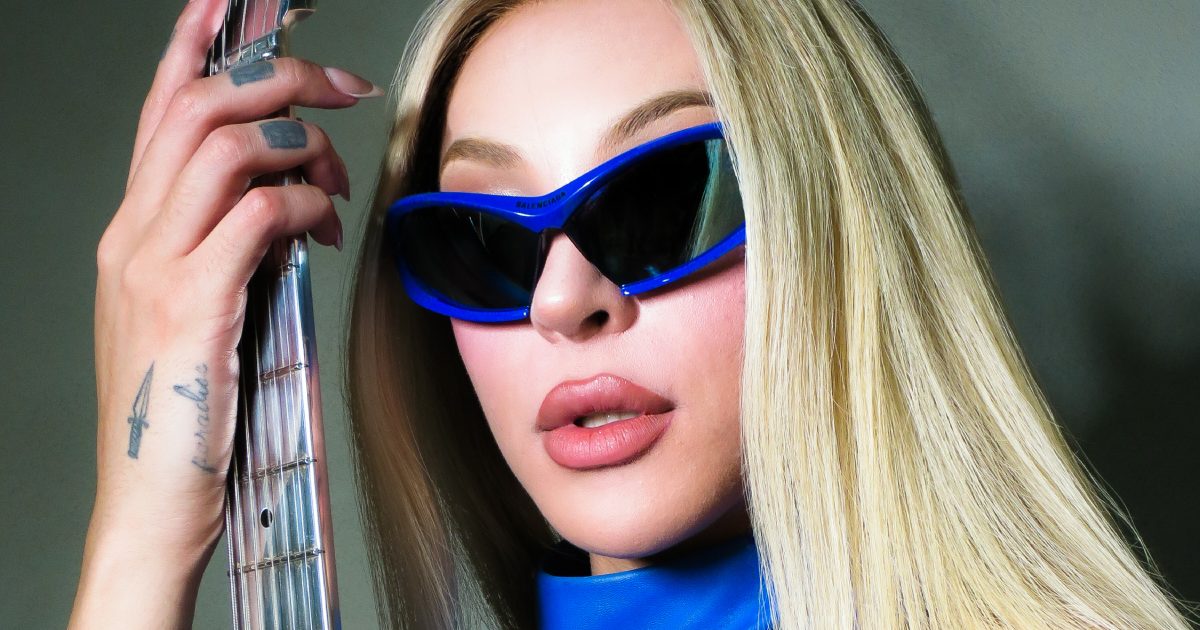 Pabllo Vittar con pelo rubio lacio, anteojos de sol azules envolventes y cuello alto azul, sosteniendo el mástil de un bajo