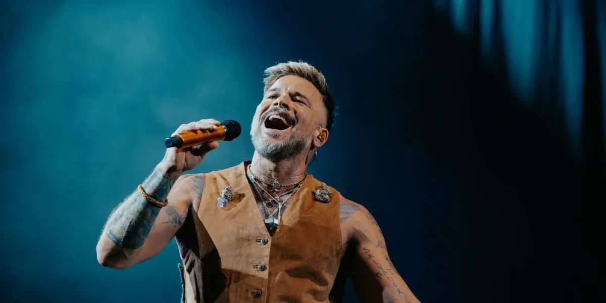 Pedro Capó en vivo: el cantante, con chaleco de cuero marrón abierto y collares, canta con los ojos cerrados y el micrófono en alto bajo una luz azul (foto de prensa)
