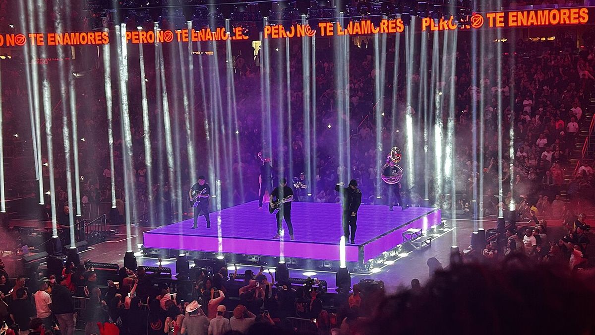Foto de archivo de Fuerza Regida en vivo en el Capital One Arena de Washington, en octubre de 2024: la banda toca sobre una plataforma iluminada de violeta en el centro del estadio, rodeada de columnas de luz blanca y con el público alrededor. Foto: Floppykart, Wikimedia Commons, CC BY 2.0