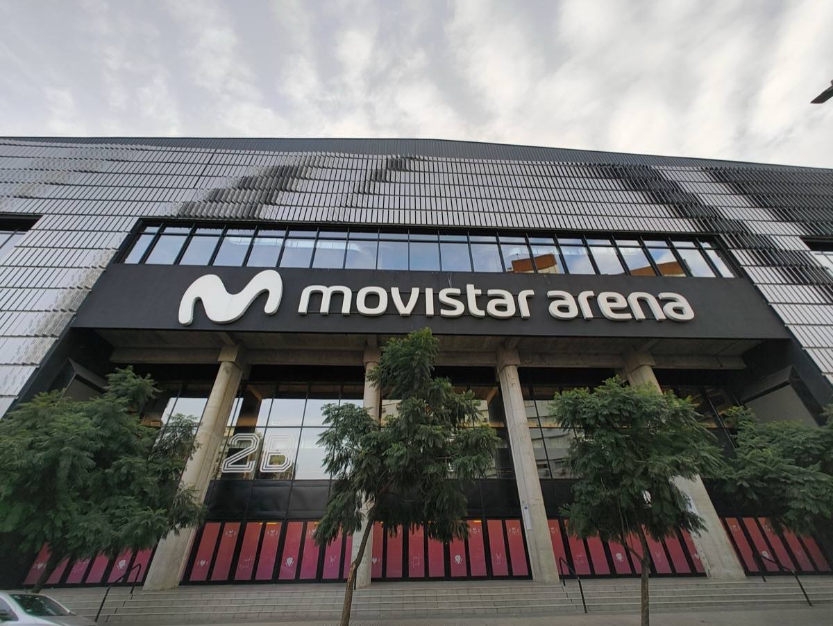 El frente del Movistar Arena de Buenos Aires, con el cartel del estadio sobre la entrada vidriada y árboles en la vereda (imagen del lugar del show). Foto: BugWarp, CC BY-SA 4.0, vía Wikimedia Commons