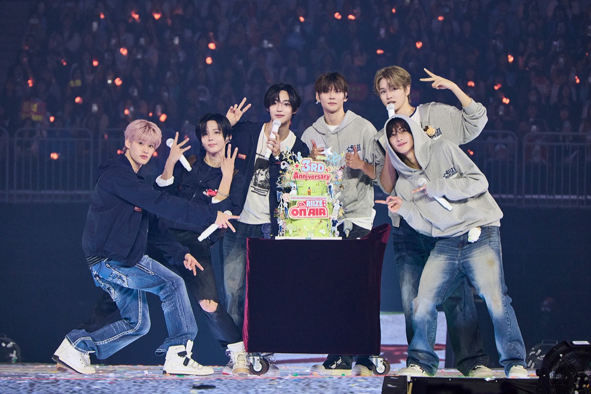 Foto oficial de archivo de RIIZE en el cierre de su fan meeting de tercer aniversario: los seis integrantes, con buzos grises, camperas y jeans, posan con micrófonos y haciendo la V con los dedos junto a una torta verde con el cartel