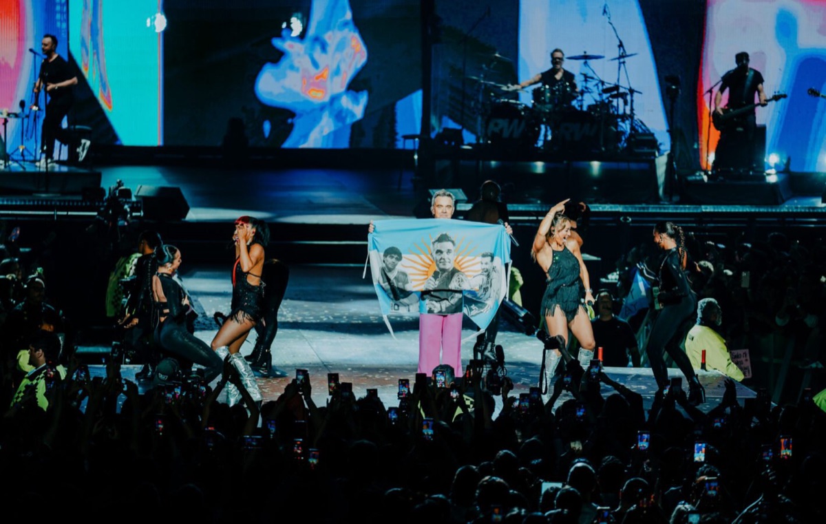 Robbie Williams con pantalón rosa sostiene una bandera argentina con su cara y el sol estampados, rodeado de bailarinas, con la banda tocando detrás y el público con celulares en primer plano