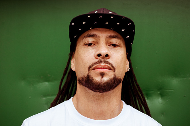 Roni Size en una foto de prensa de archivo: primer plano con gorra negra con estampado de estrellas y remera blanca, frente a una pared verde
