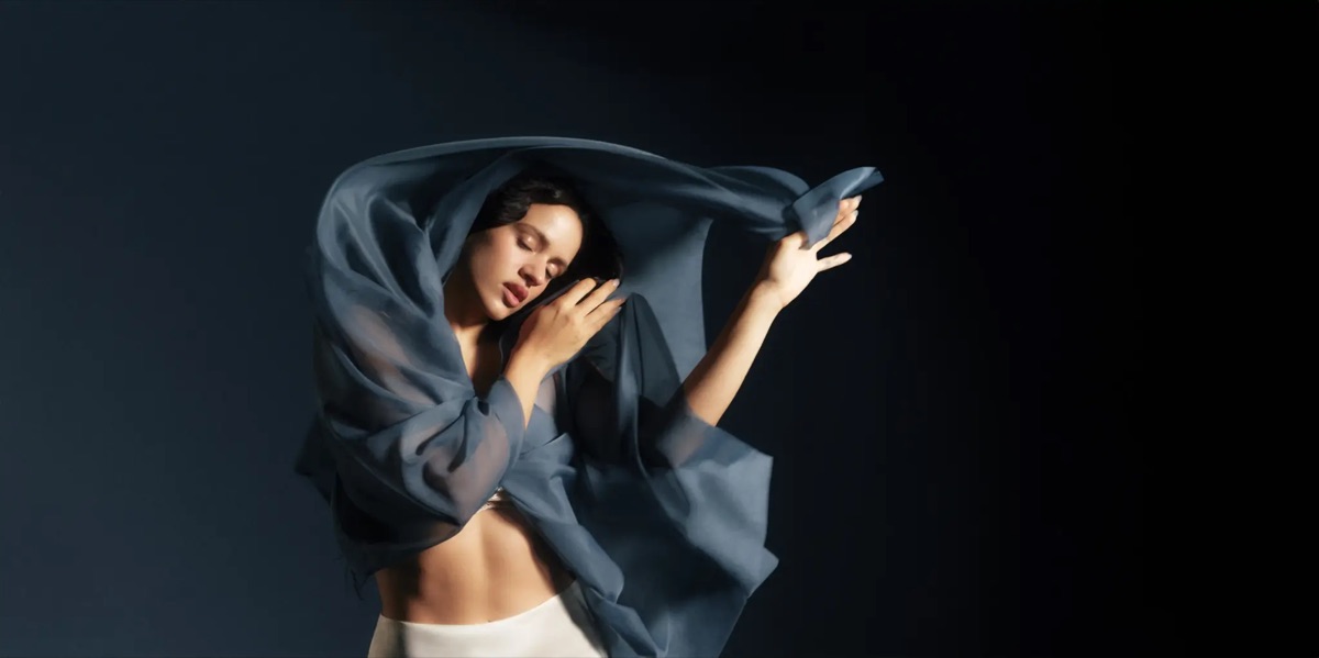 Rosalía con los ojos cerrados, envuelta en una tela de gasa azul que levanta con un brazo sobre la cabeza, con pantalón blanco y fondo azul oscuro