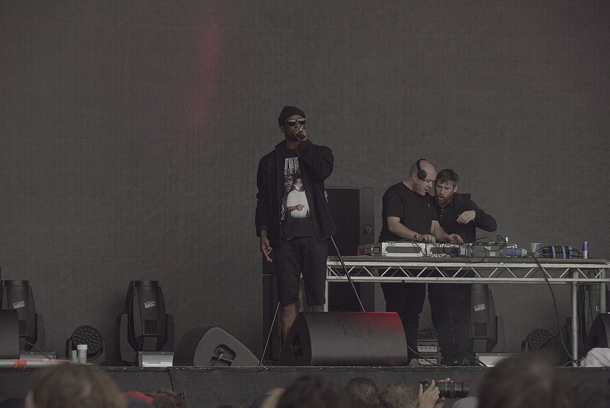 Foto de archivo de Skepta en el escenario principal del festival Field Day de Londres, en junio de 2016: canta con el micrófono en la mano, de negro y con gorra, junto a una mesa de DJ con dos personas detrás. Foto: Jwslubbock, Wikimedia Commons, CC BY-SA 4.0