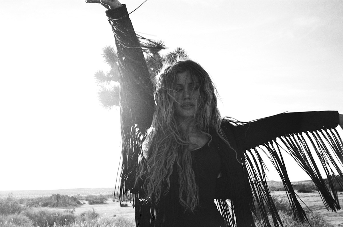 Foto de prensa oficial de Slayyyter en blanco y negro: con una campera de flecos y el pelo largo al viento, levanta un brazo en medio del desierto, con un árbol de Josué detrás y el sol a contraluz