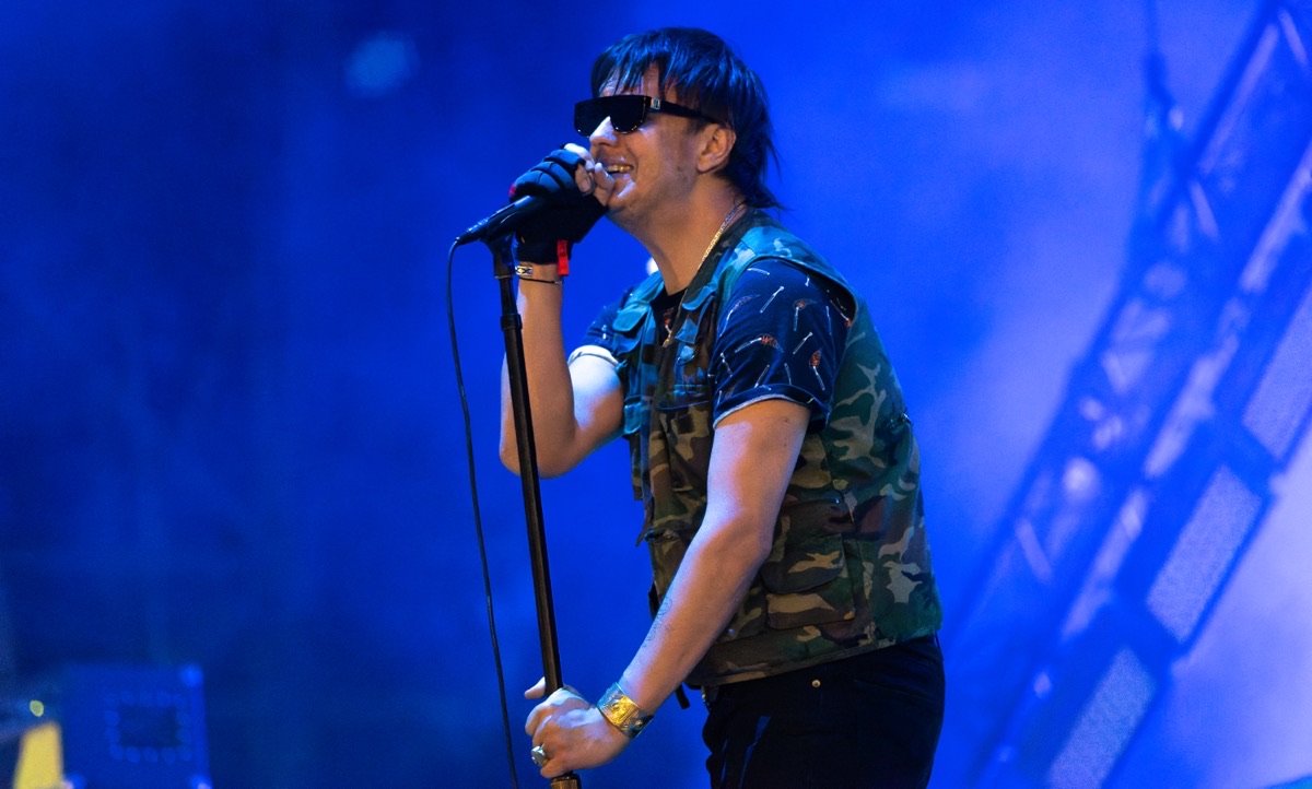 Julian Casablancas, cantante de The Strokes, en vivo en el festival NOS Alive de Lisboa en julio de 2022: canta con anteojos oscuros y camisa de camuflaje, agarrado al pie del micrófono, frente a luces azules (foto de archivo). Foto: Raph_PH, CC BY 2.0, vía Wikimedia Commons