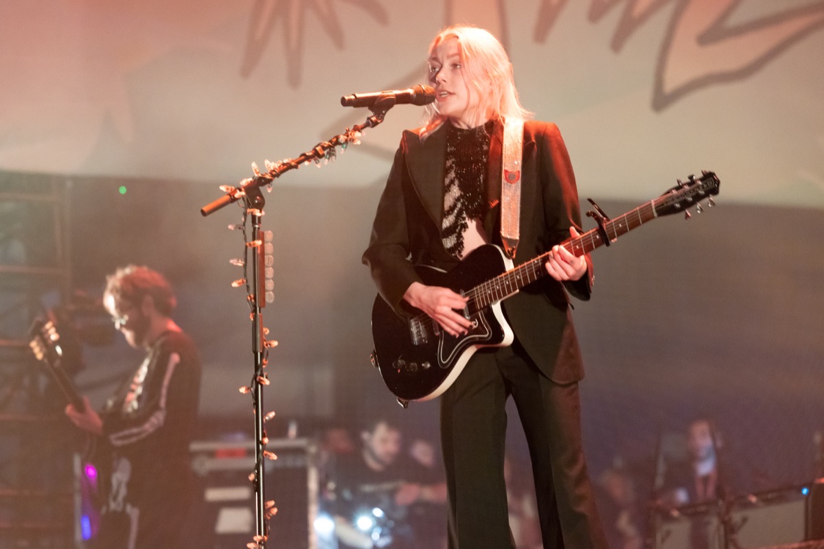Phoebe Bridgers en vivo en el festival Laneway de Sídney en febrero de 2023: canta con pelo rubio platinado, traje oscuro y una guitarra negra, frente al micrófono (foto de archivo). Foto: Bruce Baker, CC BY 2.0, vía Wikimedia Commons