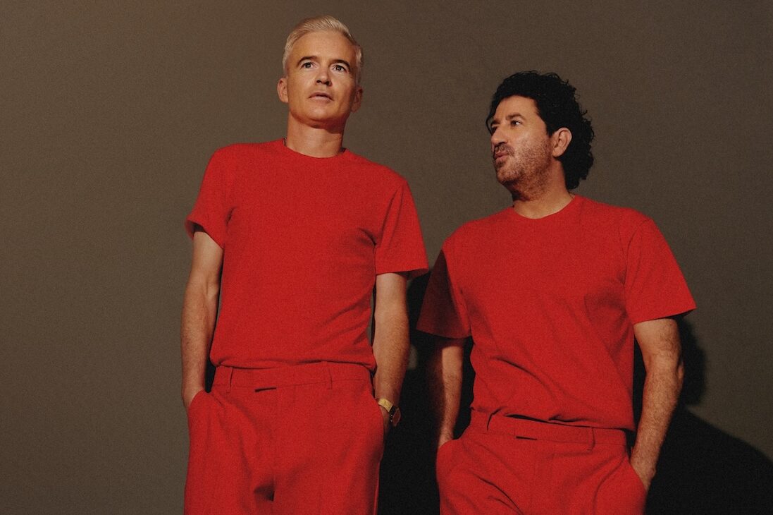 Dos integrantes de The Avalanches vestidos de rojo, con remera y pantalón, posando contra una pared marrón en una foto de prensa de la gira