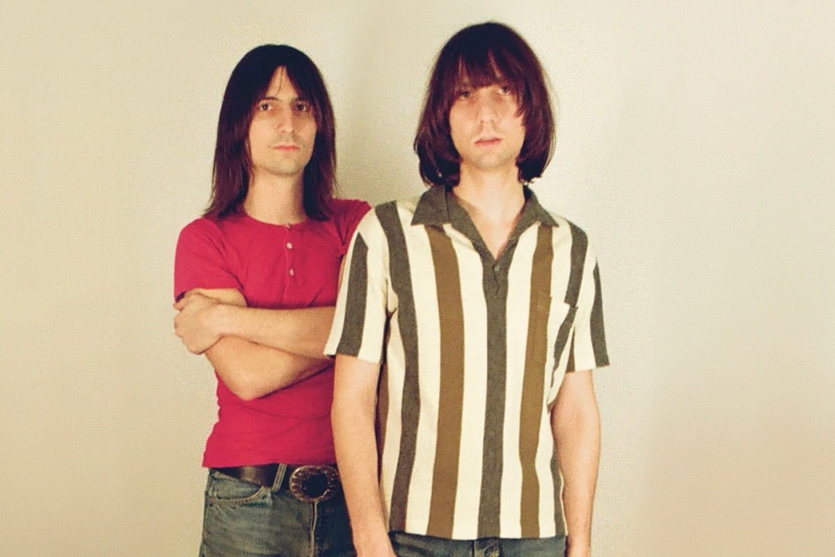 Retrato de prensa de The Lemon Twigs: los hermanos D'Addario de pelo largo sobre un fondo claro, uno con remera roja y los brazos cruzados, el otro adelante con camisa a rayas verticales marrones y blancas