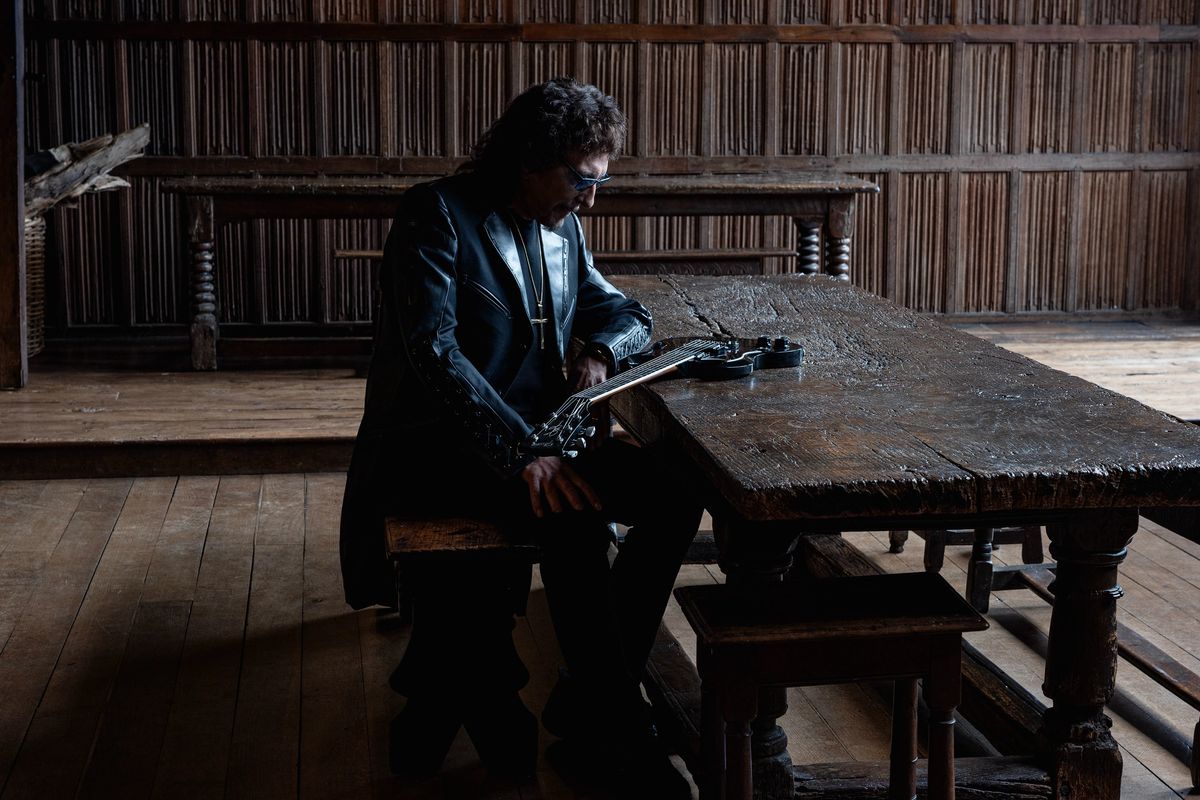 Foto de prensa oficial de Tony Iommi para «From the Dark»: con campera de cuero negra y anteojos, sentado a una mesa larga de madera en un salón antiguo con paredes de madera, sostiene una guitarra eléctrica