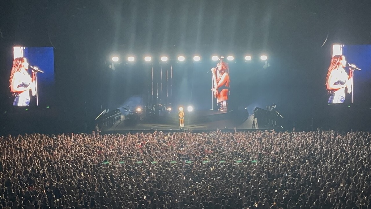 Foto de archivo de Tate McRae en el escenario durante un show en Adelaida, Australia, en noviembre de 2024: la cantante aparece en las pantallas gigantes a los costados mientras el estadio lleno la mira bajo una fila de reflectores blancos. Foto: Avatar5991, Wikimedia Commons, CC BY-SA 4.0