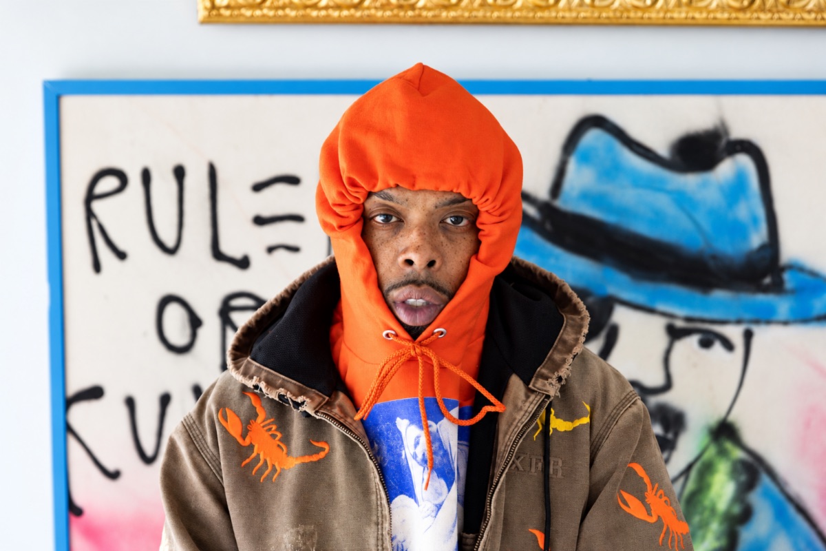 Foto de prensa de Westside Gunn con capucha naranja y campera marrón con escorpiones bordados, frente a una pintura con grafitis