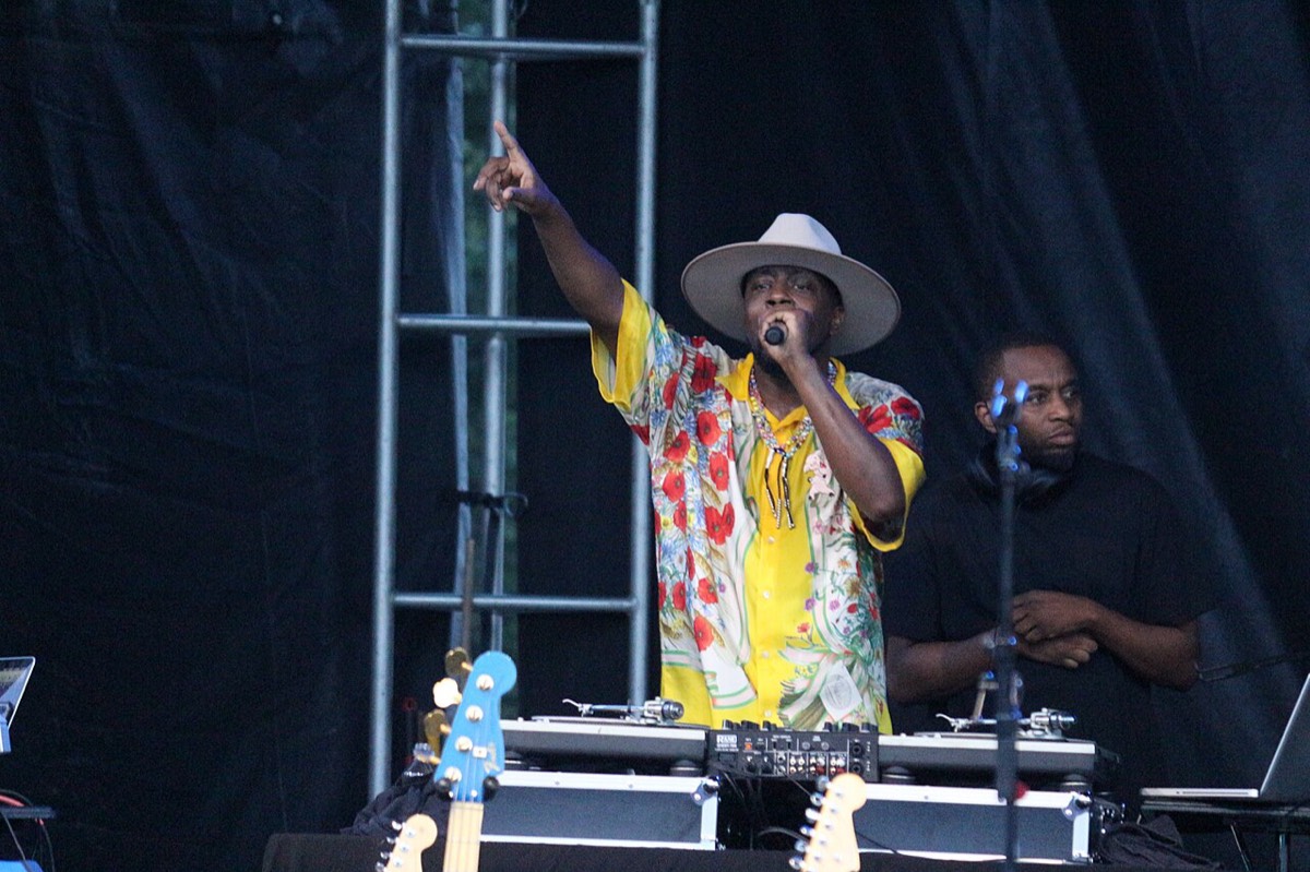 Wyclef Jean en vivo en el Memorial Park de Omaha, en agosto de 2021 (foto de archivo): con sombrero blanco de ala ancha y camisa amarilla con flores rojas, señala hacia arriba con el micrófono en la mano detrás de una consola de DJ. Foto: Matt Johnson, Wikimedia Commons, CC BY 2.0