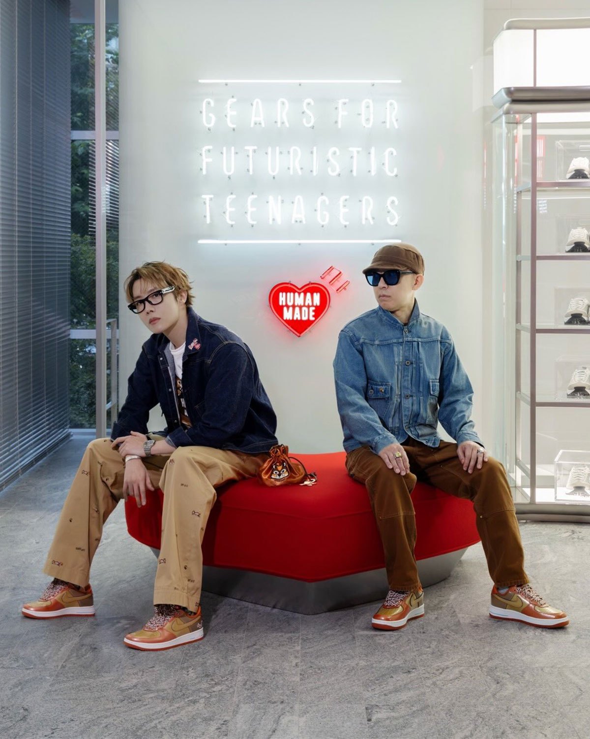 j-hope y NIGO sentados en un banco rojo frente a un cartel de neón de Human Made, los dos con el Nike Air Force 1 '01 Burnt Sunrise en charol cobre y dorado