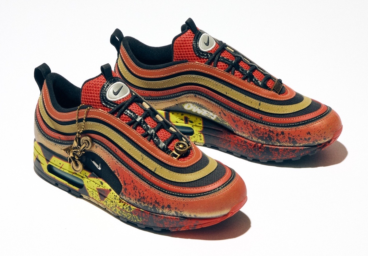 Nike Air Max 1/97 de la colección Doernbecher Freestyle 22 sobre fondo blanco, con las líneas onduladas del Air Max 97 en rojo, dorado y negro, salpicaduras oscuras, una zona amarilla en el talón y un colgante dorado en los cordones