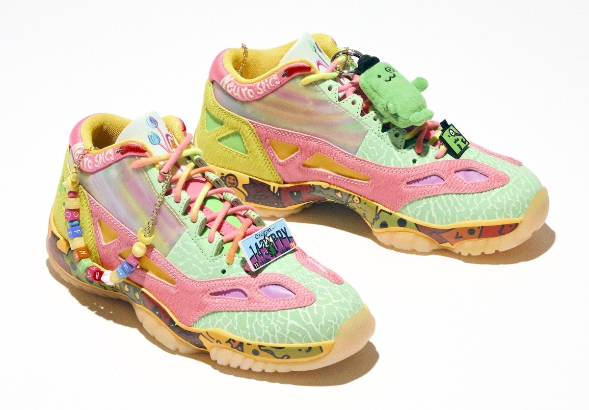 Air Jordan 11 Low IE de la colección Doernbecher Freestyle 22 sobre fondo blanco, con upper verde agua con textura, paneles rosas, zonas tornasoladas, suela translúcida, cordones con cuentas de colores y un muñeco de peluche verde colgado del cuello