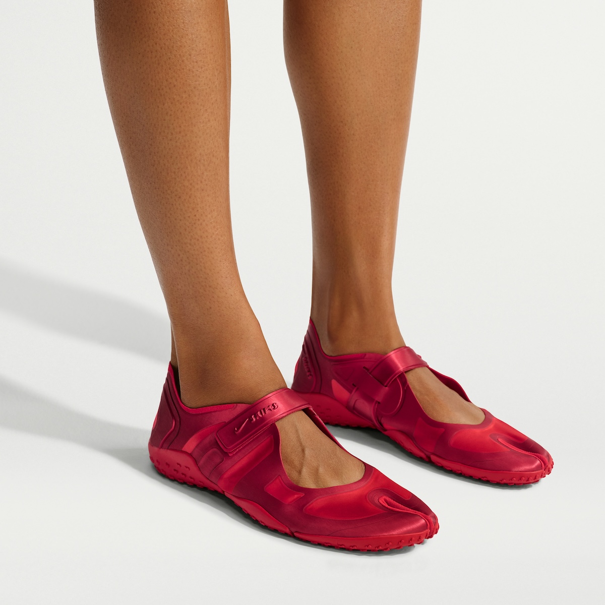 Pies con la NikeSKIMS Rift Satin University Red puesta sobre fondo blanco, con la punta dividida, la tira de abrojo sobre el empeine y la suela roja