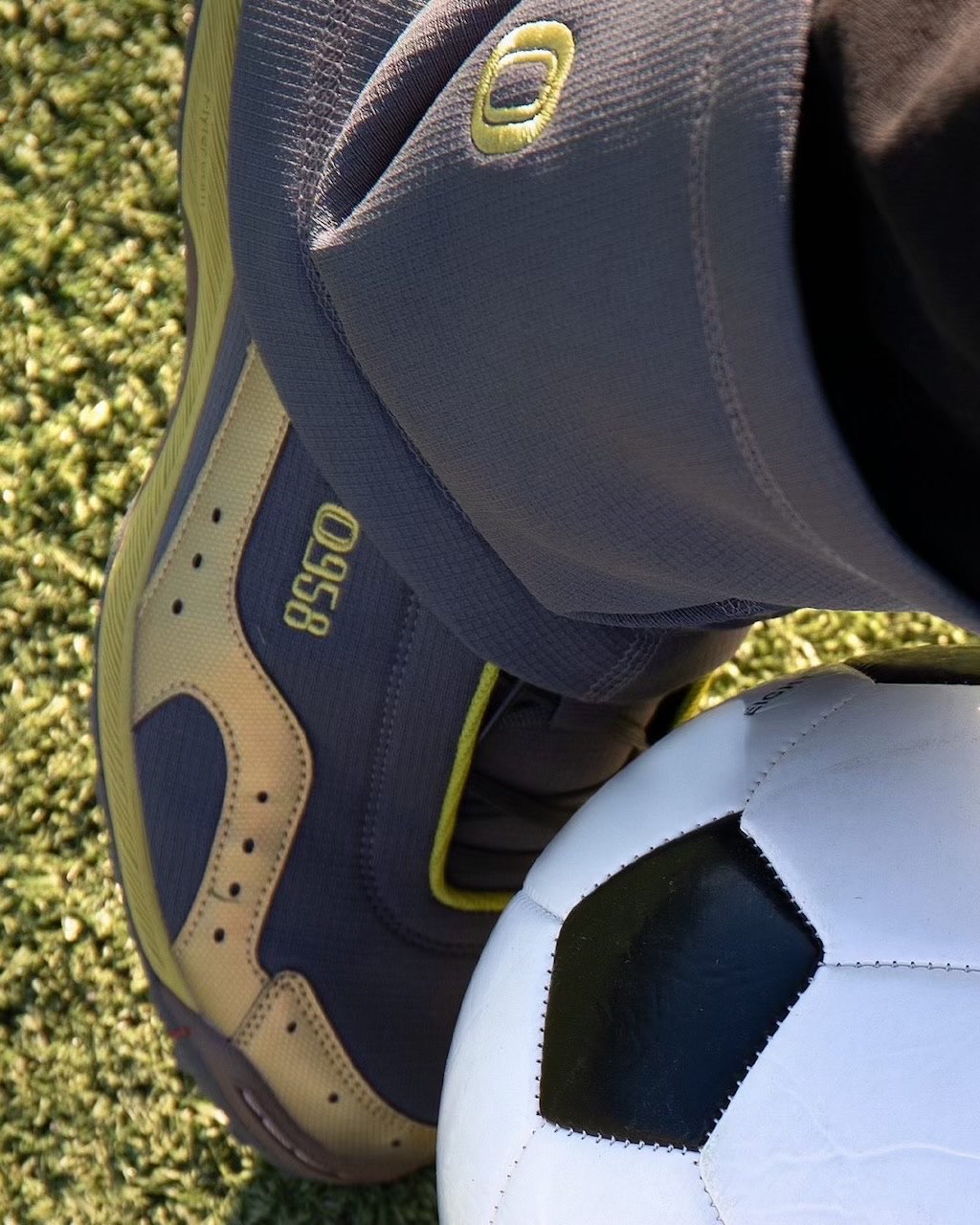 Detalle de la ASICS GEL-BELARRAOTTO de OTTO 958 con el pie apoyado sobre una pelota de fútbol, con la capellada gris oscura, el logo OTTO 958 en el lateral, detalles dorados y la polaina gris encima