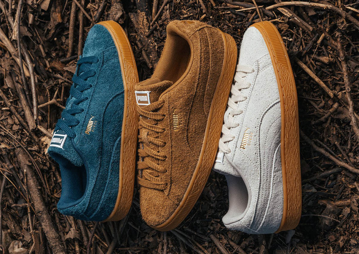 Tres PUMA Suede Forever sobre ramas secas: Midnight Petrol azul petróleo, Kona Brown marrón y Silver Fog gris claro, todos con gamuza de pelo largo, logo dorado y suela gum