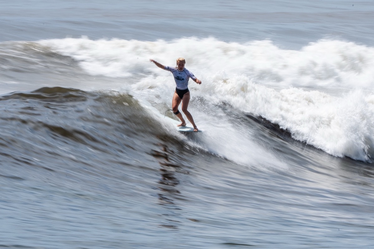 Thilde Rasmussen, con lycra blanca, camina sobre su longboard con los brazos abiertos en una ola de El Sunzal, con la espuma detrás