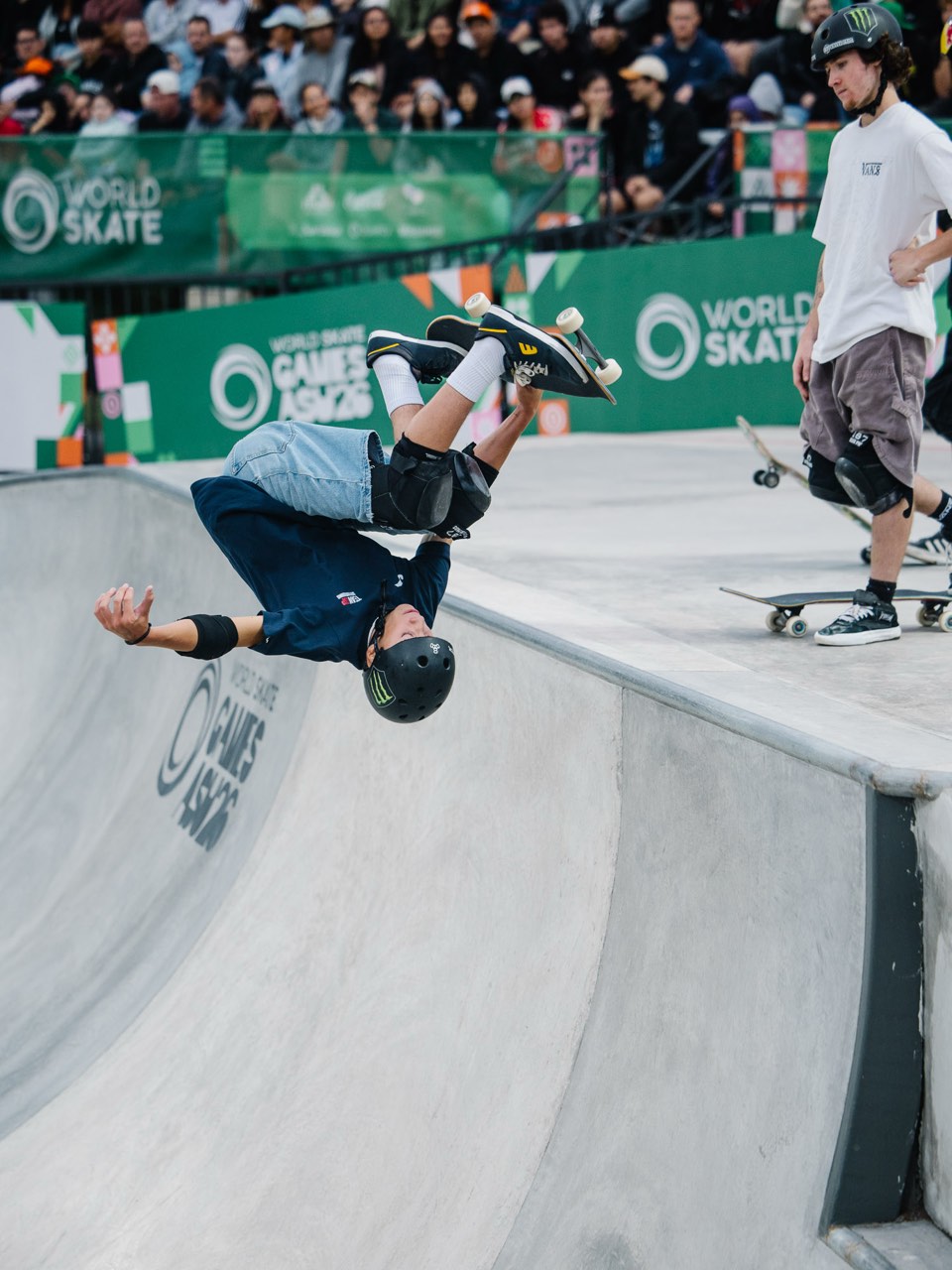 Egoitz Bijueska, con casco negro y buzo azul, gira cabeza abajo sobre el borde del bowl agarrando la tabla, con el público y los carteles de World Skate de fondo