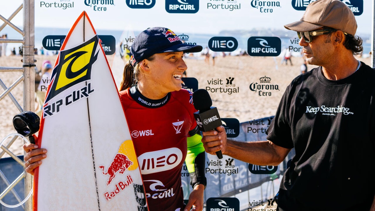 Molly Picklum, con la lycra roja del MEO Rip Curl Pro Portugal y gorra azul, sonríe con su tabla blanca mientras le hacen una entrevista en la playa de Supertubos