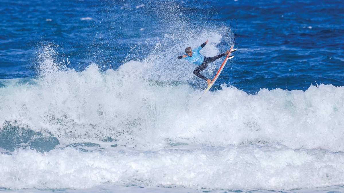 Yago Domínguez, con lycra celeste, vuela por encima de la espuma de una ola con la tabla en el aire y el mar azul detrás
