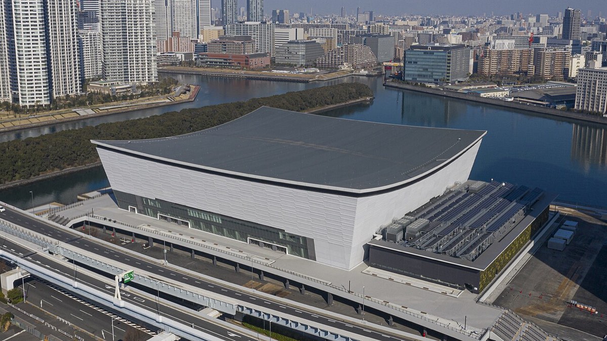 El Ariake Arena visto desde el aire, con su techo curvo gris, junto a un canal y rodeado de edificios de la bahía de Tokio