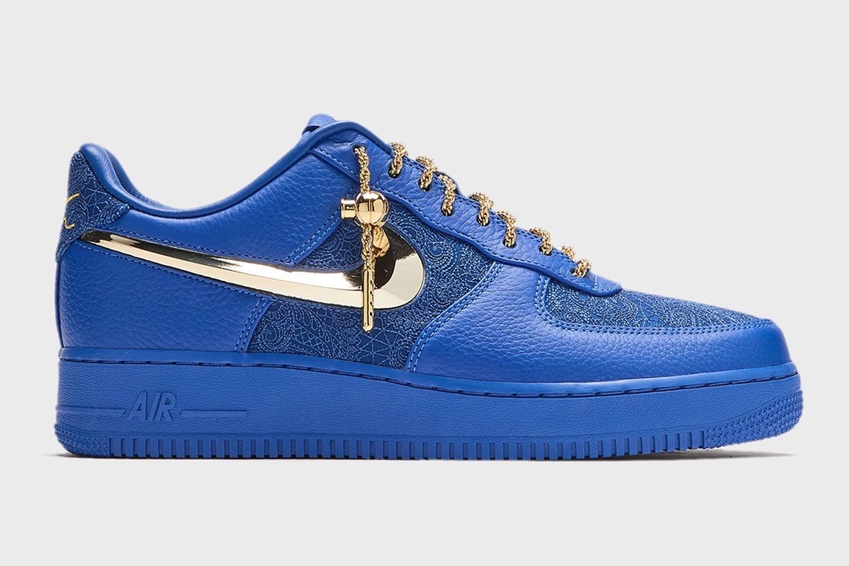 Victor Solomon x Nike Air Force 1 Low Blue Trophies de perfil sobre fondo gris claro, de cuero azul con el Swoosh dorado metálico, cordones de soga dorados y un dije colgante
