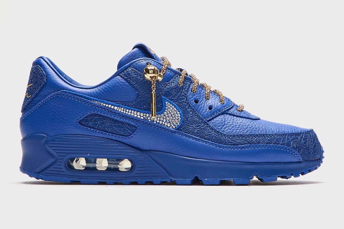 Victor Solomon x Nike Air Max 90 Blue Trophies de perfil sobre fondo gris claro, todo azul con el Swoosh brillante, cordones de soga dorados y la cámara de aire